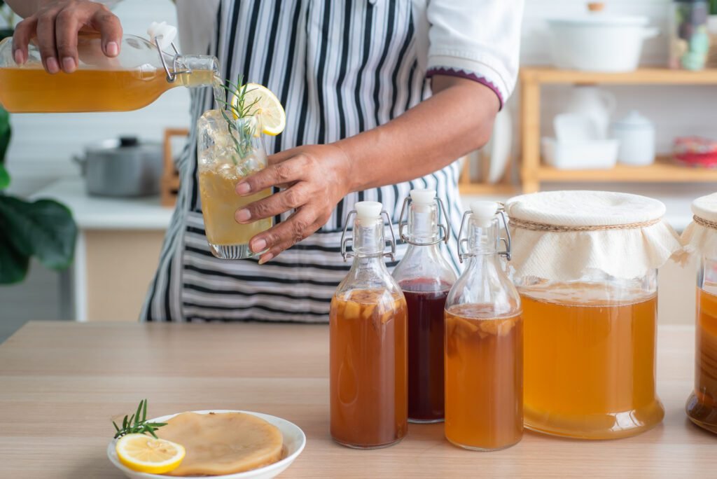 1920 chef s hand holds a bottle and a glass of homemade fermented kombucha tea variety of flavors in bottles and glass jars arranged mix with fruit scoby on wooden table healthy natural probiotic drink